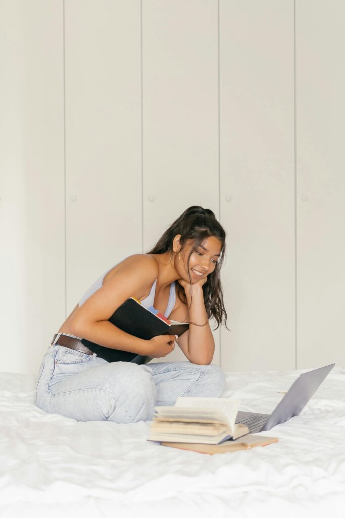 Smiling woman studying with books and laptop on bed, embracing e-learning at home.